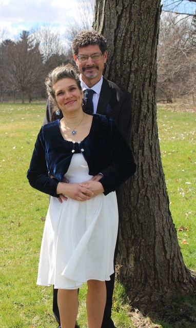 A man stands behind his wife and hugs her in front of a tree