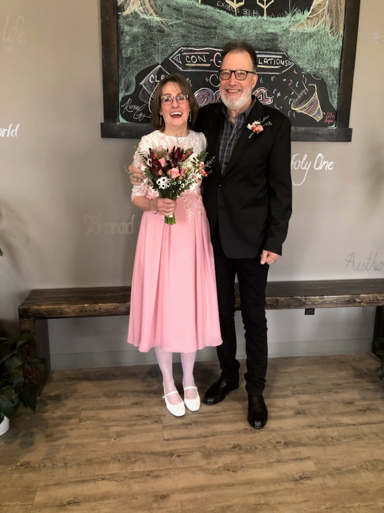 A mature Christian bride in a pink skirt holds a bouquet of flowers and laughs excitedly next to her bearded husband, dressed in a black suit, as they stand in a church lobby