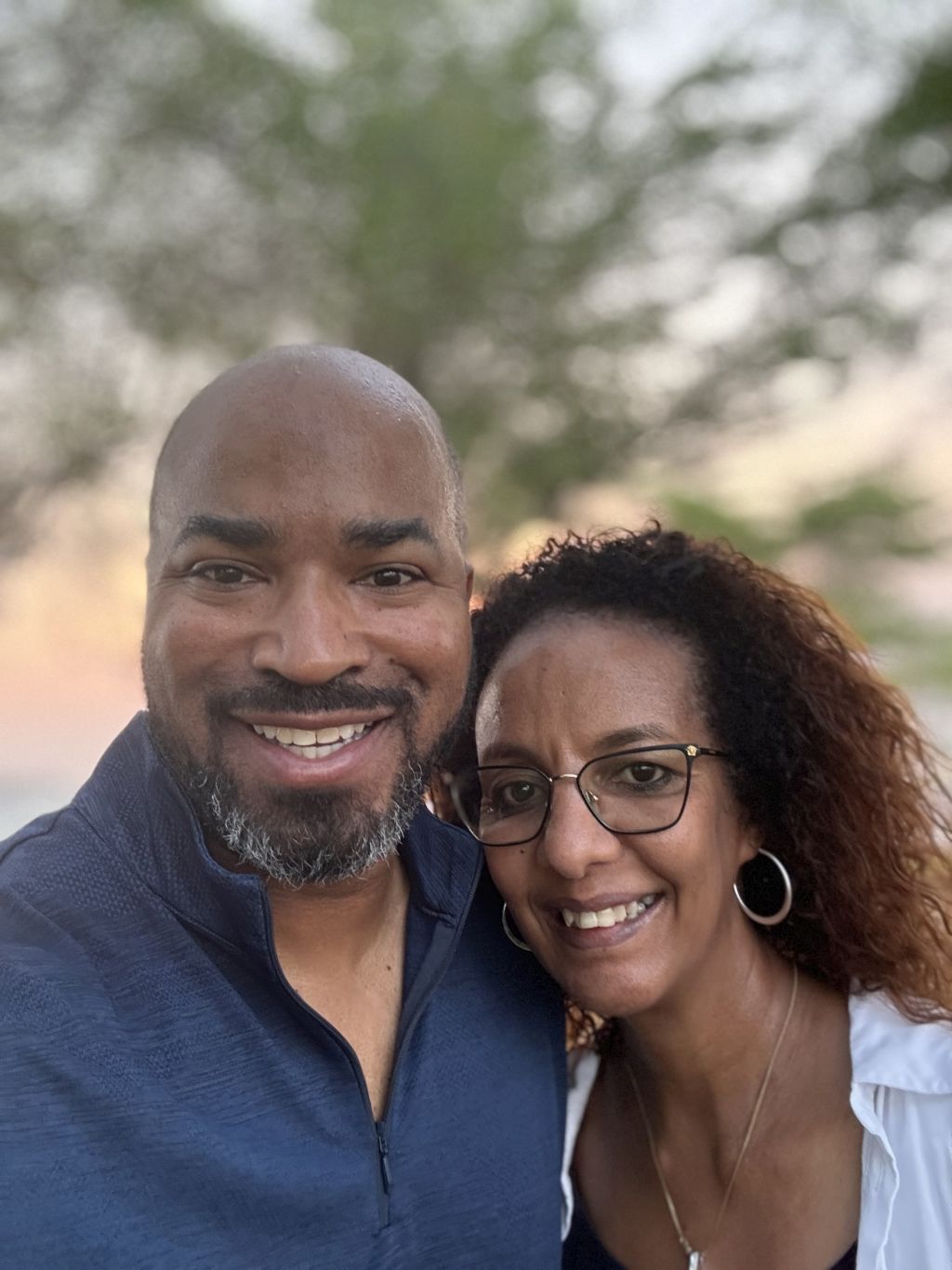 A bearded Christian man smiles with joy as his wife leans in for a selfie outdoors