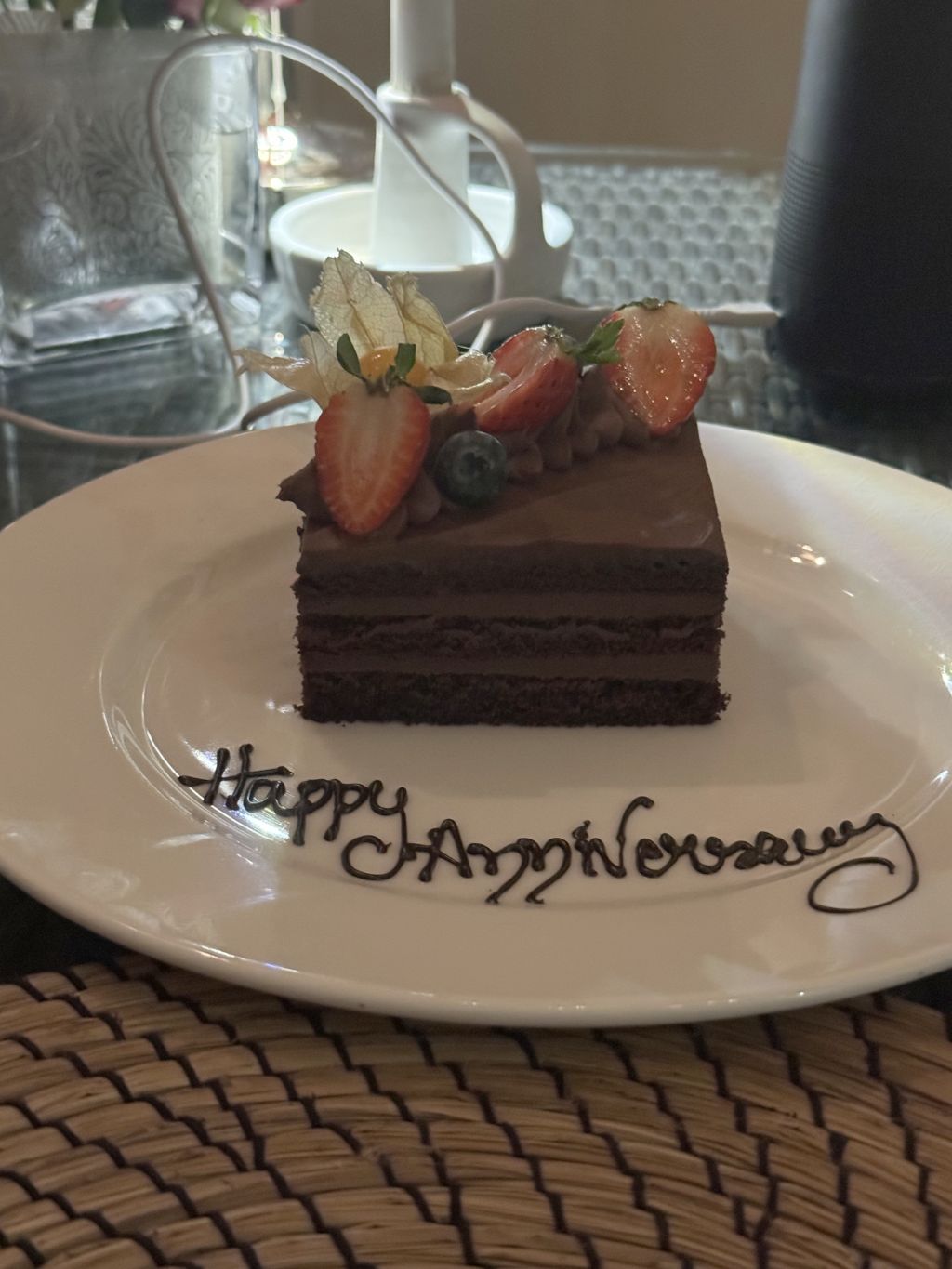 A piece of chocolate cake with strawberries and blueberries is shown on a plate with the words Happy Anniversary