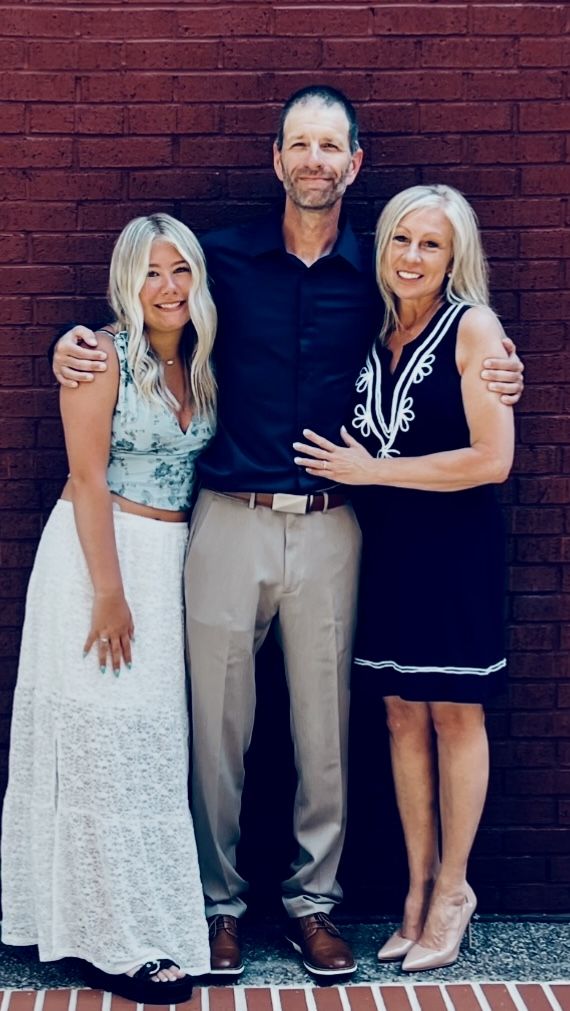 A man stands in front of a brick wall holding on to his daughter and his new wife