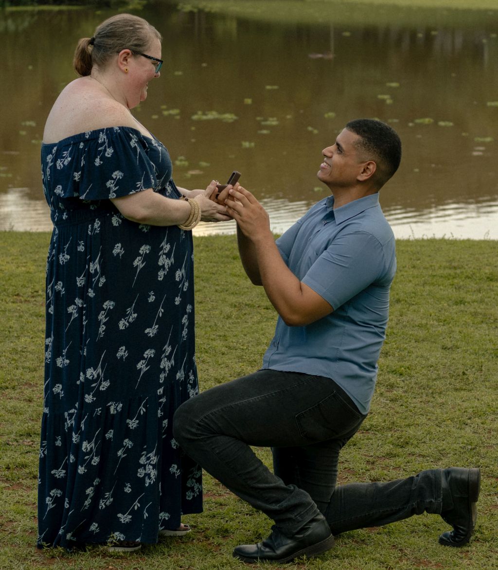 A man proposes on bended knee to a woman in a strapless dress by the water
