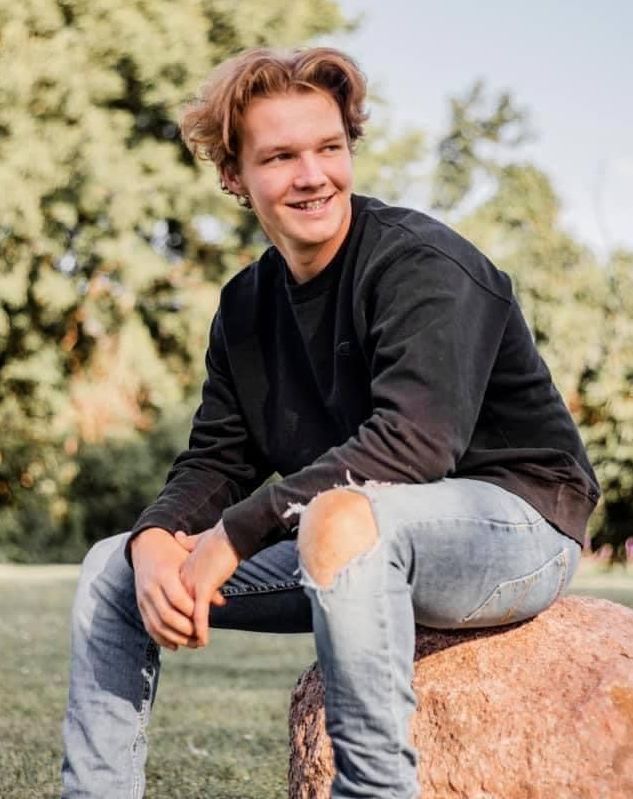 A young man with ripped jeans sits on a rock while smiling