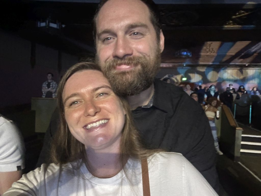 A pretty woman smiles as a bearded man leans behind her while they're at an event