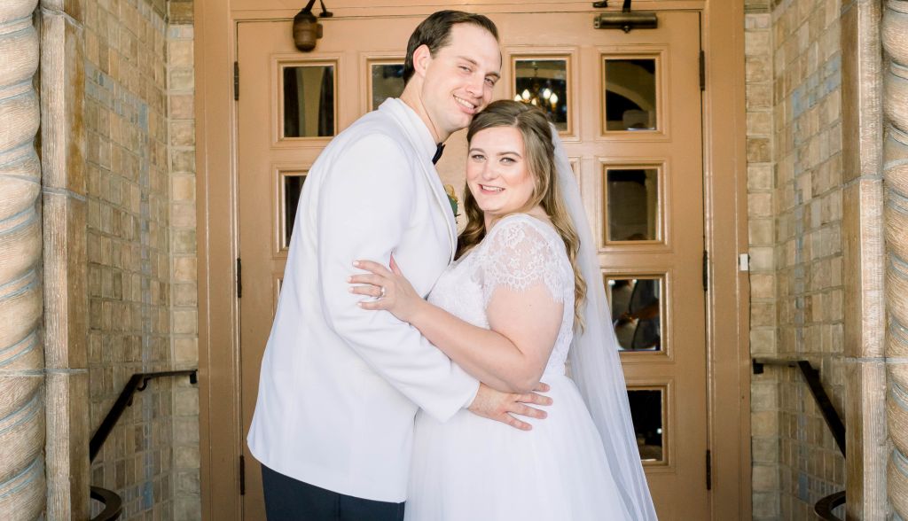 A glowing bride in white poses in a doorway with her new husband, who is in a white suit