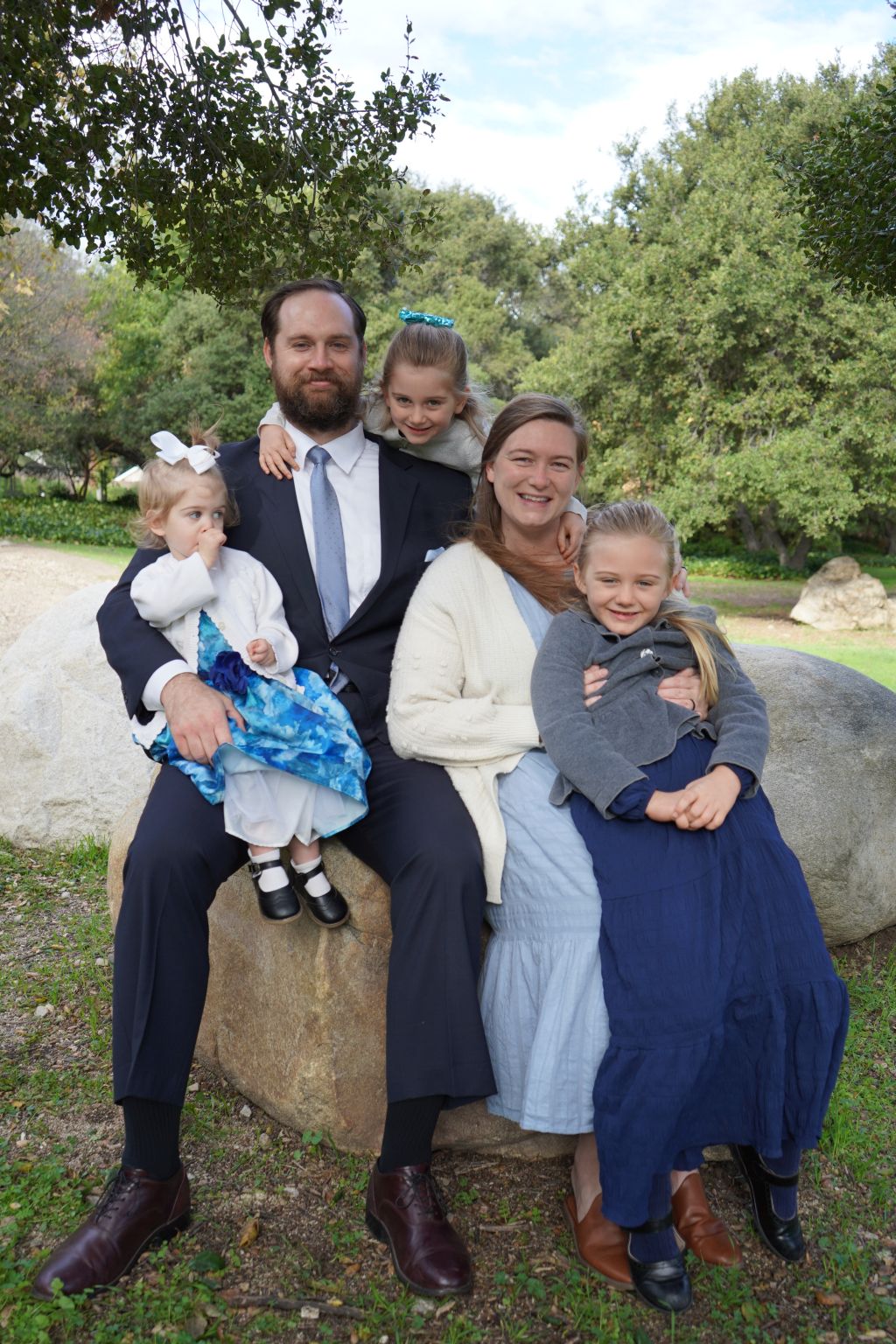 A proud Christian couple pose on a big rock in a park, with their 3 daughters