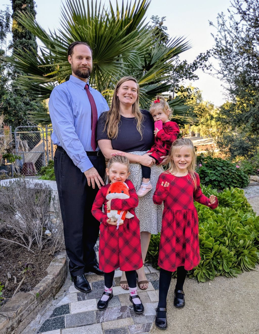 A well dressed man stands proudly with his pretty wife and their 3 daughters, all in matching checkered dresses