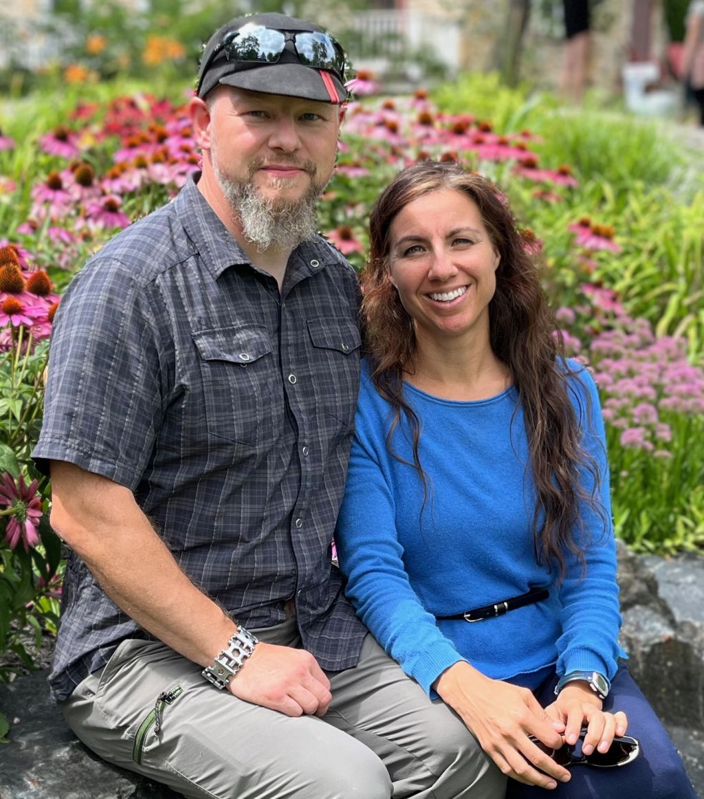 A Christian man with a beard wearing a hat and sunglasses sits next to a pretty woman in a blue top as they sit in front of flowers in the Spring