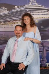 A well dressed couple stand and smile in front of a cruise ship