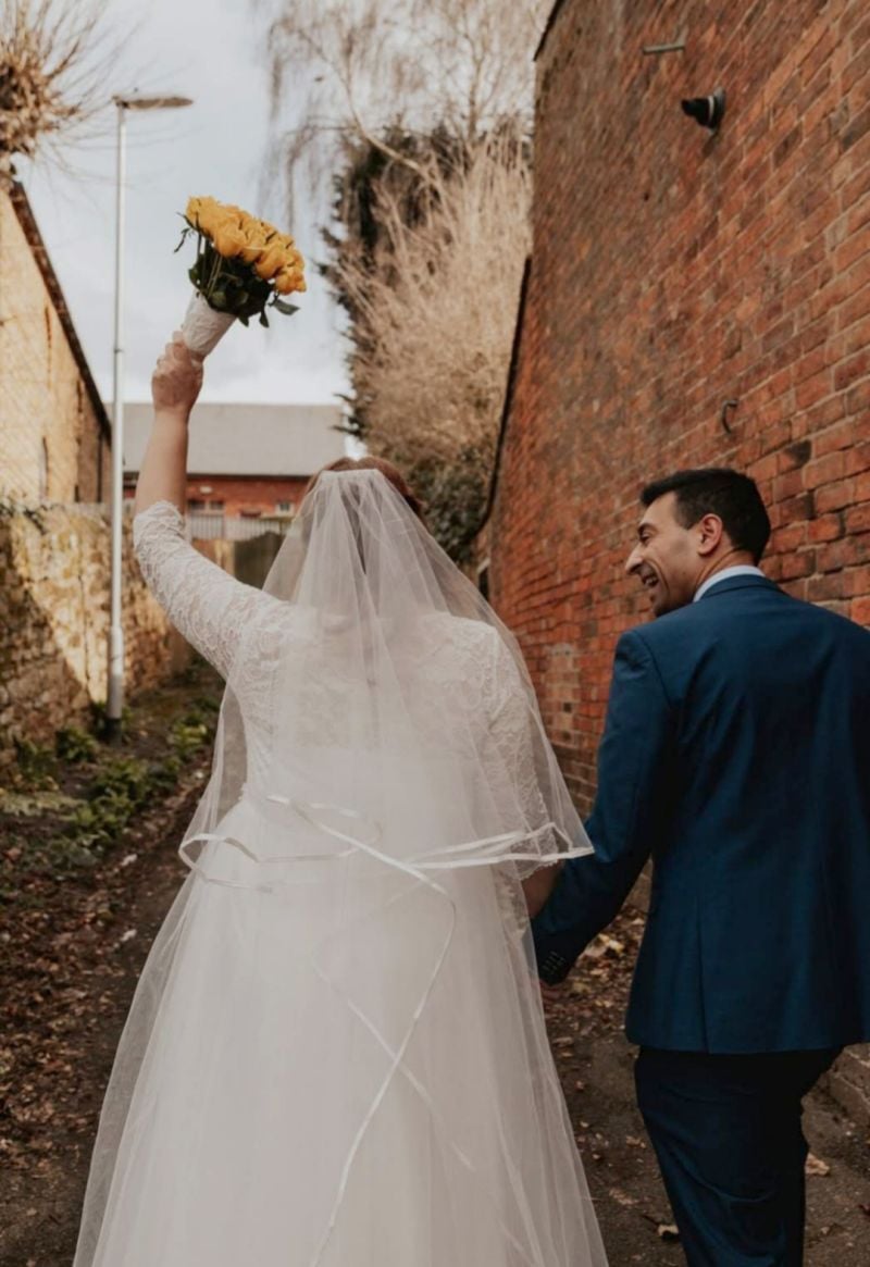 A bride and groom laugh while walking away, as she holds up her bouquet of flowers