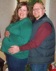 A man laughs as he puts his arms around his wife who shows off her baby bump