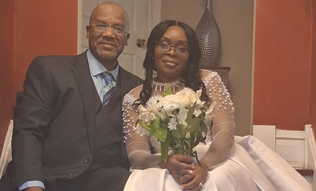 A very happy groom smiles as he sits on some stairs and leans into his bride, who looks radiant holding a white bouquet of flowers